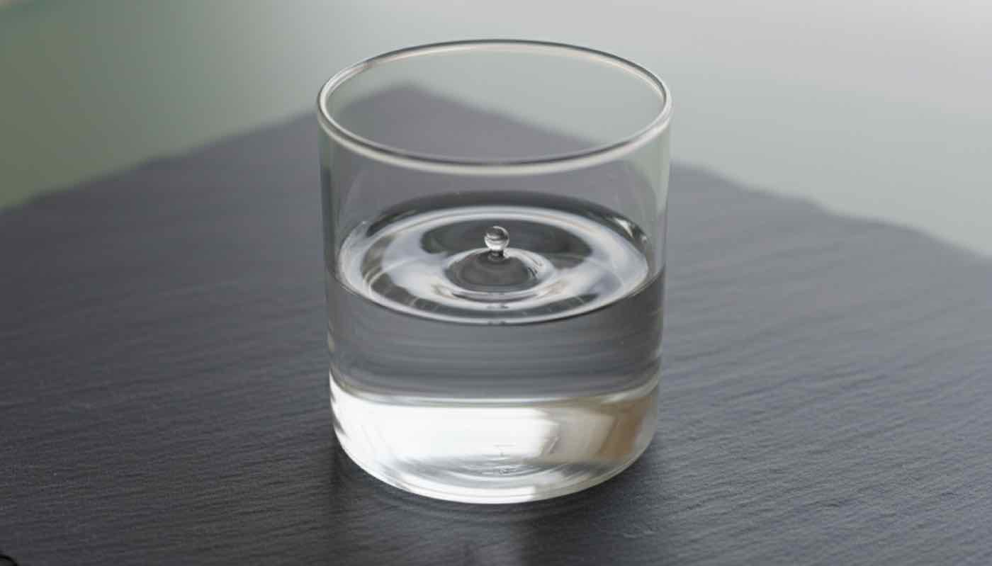 Top-down close-up of clean transparent water in a glass beaker on a slate surface with a single ripple, calm investigative editorial composition.